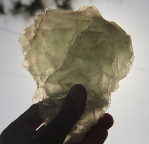 A hand displays Bahgsu Jewels’ Raw Mongolian Fluorite (cosmic city) against the sky, revealing its thin, plate-like form and soft yellow-green hue—an exquisite specimen cherished for aura cleansing.