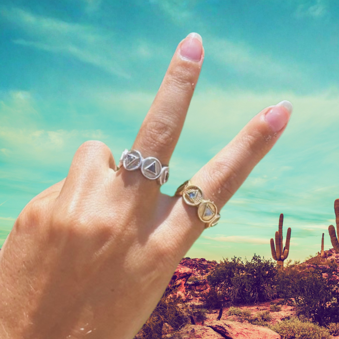 Hand with two rings against a desert landscape with cacti and blue sky.